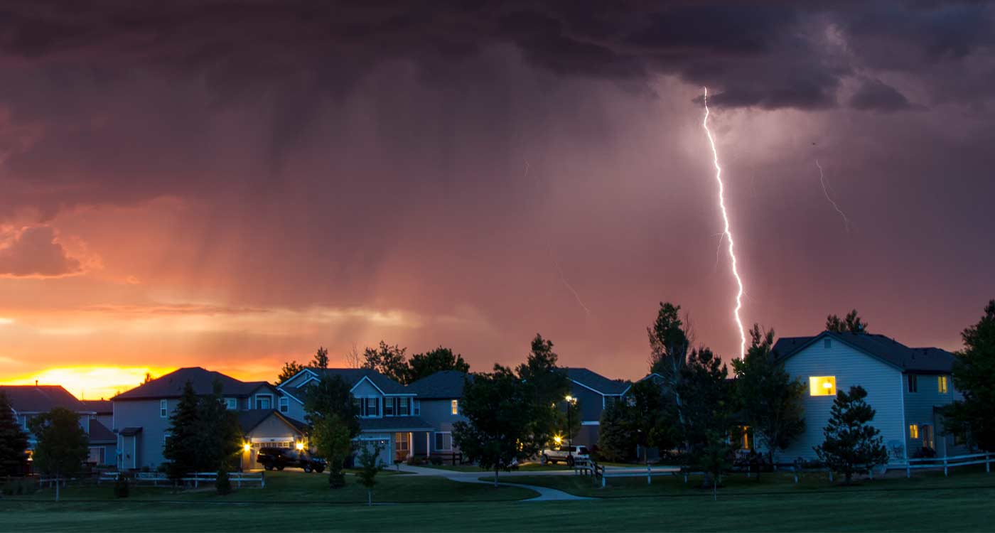 Lightning strikes beyond a suburban neighborhood under dark storm clouds at dusk
